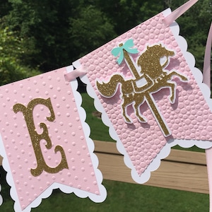 May include: Pink and white banner with gold glitter letters spelling "E" and a gold glitter carousel horse with a turquoise bow. The banner has a scalloped edge and a pink polka dot pattern.