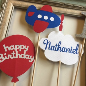 May include: Four cake toppers for a first birthday party. The toppers are a red balloon with the words "happy birthday" in white, a blue airplane with red accents, a white cloud with the name "Nathaniel" in blue, and a silver glitter number one.