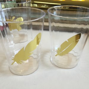 Puede incluir: Dos vasos de pl&aacute;stico transparentes con calcoman&iacute;as de plumas doradas. Las plumas son un dise&ntilde;o simple de silueta.