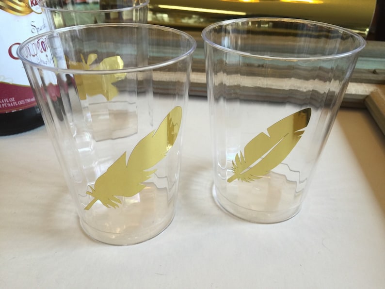 Puede incluir: Dos vasos de pl&aacute;stico transparentes con calcoman&iacute;as de plumas doradas. Las plumas son un dise&ntilde;o simple de silueta.