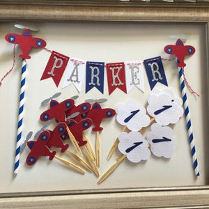 Puede incluir: Toppers para pastel con tem&aacute;tica de avi&oacute;n en rojo, blanco y azul con una pancarta que dice "PARKER". El conjunto incluye 10 toppers de avi&oacute;n, 4 toppers de nube y una pancarta.