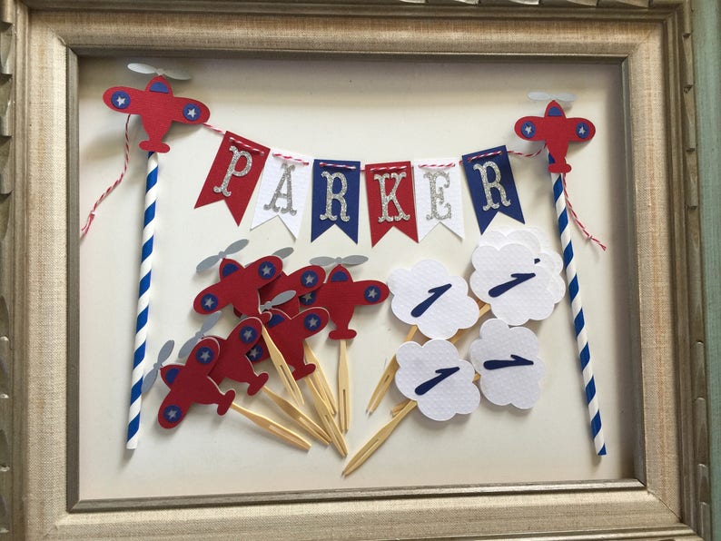 Puede incluir: Toppers para pastel con tem&aacute;tica de avi&oacute;n en rojo, blanco y azul con una pancarta que dice "PARKER". El conjunto incluye 10 toppers de avi&oacute;n, 4 toppers de nube y una pancarta.