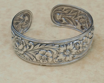 May include: A silver bangle bracelet with an intricate floral design. The bracelet is made of metal and has a smooth, polished finish.