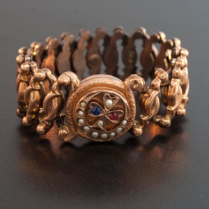 May include: A gold bracelet with a clover-shaped charm set with pearls, a blue stone, and a red stone.