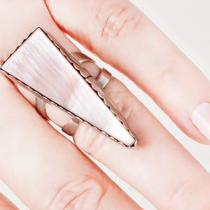 May include: A large, silver-toned ring with a rectangular, iridescent, mother-of-pearl inlay. The ring has a geometric design with a series of small, connected silver bands. The ring is worn on a finger against a light background.