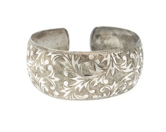 May include: Silver-toned cuff bracelet with an open design. The wide band features an intricate floral pattern etched across the surface. The bracelet has a slightly aged appearance, with visible wear and tear.