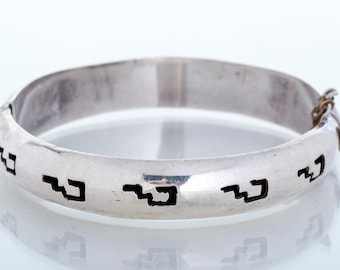 May include: A silver bangle bracelet with a hinged clasp and a repeating geometric pattern etched into the surface.