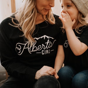 K&ouml;nnte beinhalten: Eine Frau und ein junges M&auml;dchen tragen passende schwarze T-Shirts mit wei&szlig;em Text. Das Shirt der Frau sagt "Alberta Girl" mit einer Berggrafik. Das Shirt des M&auml;dchens sagt "Girl".