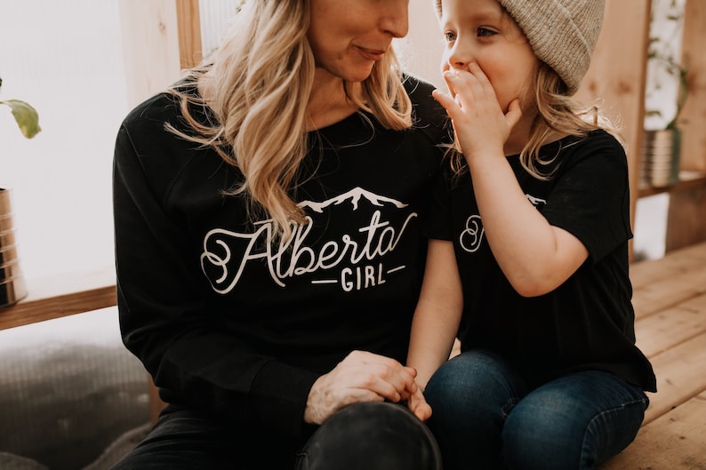 K&ouml;nnte beinhalten: Eine Frau und ein junges M&auml;dchen tragen passende schwarze T-Shirts mit wei&szlig;em Text. Das Shirt der Frau sagt "Alberta Girl" mit einer Berggrafik. Das Shirt des M&auml;dchens sagt "Girl".