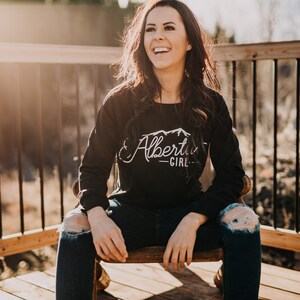 K&ouml;nnte beinhalten: Eine Frau tr&auml;gt ein schwarzes Langarmshirt mit einem wei&szlig;en Aufdruck, der "Alberta Girl" und ein Bergpanorama zeigt. Sie sitzt auf einem Holzstuhl mit gekreuzten Beinen und tr&auml;gt eine blaue Jeans mit L&ouml;chern in den Knien.