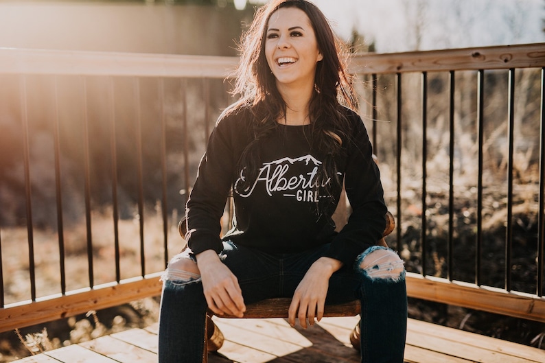 K&ouml;nnte beinhalten: Eine Frau tr&auml;gt ein schwarzes Langarmshirt mit einem wei&szlig;en Aufdruck, der "Alberta Girl" und ein Bergpanorama zeigt. Sie sitzt auf einem Holzstuhl mit gekreuzten Beinen und tr&auml;gt eine blaue Jeans mit L&ouml;chern in den Knien.