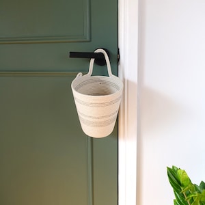May include: A small, off-white rope basket with grey horizontal stripes hangs from a black door handle. The basket has a cylindrical shape with a looped handle. The door is painted a muted green, and the wall is white.