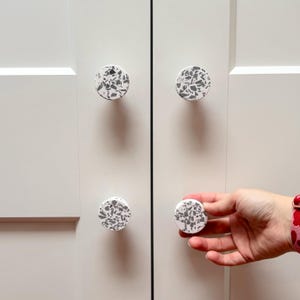 May include: Four circular cabinet knobs with a white base and a speckled grey terrazzo pattern. The knobs are mounted on a pair of light cream-coloured cabinet doors, with one knob being held by a hand to demonstrate its size and shape.