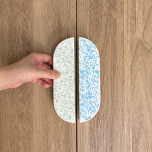 May include: Two semi-circular cabinet handles featuring a terrazzo pattern. One handle displays a white base with light green flecks, while the other features a white base with bright blue flecks. Both handles have a smooth, rounded edge design.