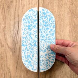 May include: Two semi-circular cabinet handles featuring a white base with a scattered light blue terrazzo pattern. Each handle has a smooth, rounded top edge and a flat vertical side, designed to be placed together to form a single oval shape.
