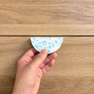 May include: A semi-circular cabinet handle featuring a white base with a speckled terrazzo pattern in bright light blue. The hardware has a smooth, flat surface and a curved edge, designed for mounting onto furniture or cabinetry doors and drawers.