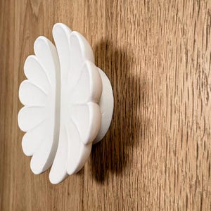 May include: A matte white cabinet knob shaped like a flower with multiple rounded petals. The knob features a circular base and a dimensional, layered design that extends outward, creating a distinct floral silhouette against the surface.