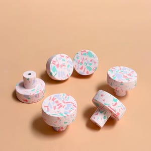 May include: A set of five round cabinet knobs featuring a light pink base with a speckled terrazzo pattern in shades of mint green, coral pink, and lavender. Each knob has a flat circular face and a cylindrical base with a central metal screw hole.