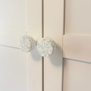 May include: Two circular cabinet knobs attached to white cabinet doors. Each knob features a white base with a speckled terrazzo pattern consisting of light blue-grey geometric fragments embedded across the flat, round front surface.