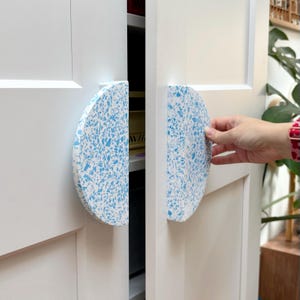 May include: Two semi-circular cabinet handles attached to white double doors. The handles feature a white base with a speckled pattern of light blue, angular fragments, creating a terrazzo-like appearance on the curved, flat-faced hardware.