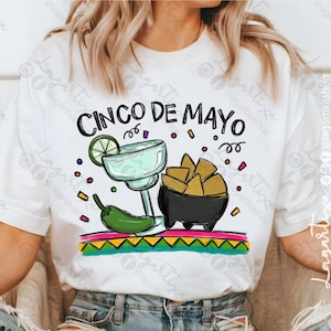 May include: White t-shirt with a colorful graphic design that says "Cinco de Mayo". The design includes a margarita glass, a bowl of chips, a lime wedge, and a green chili pepper.