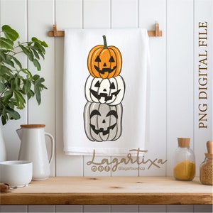 Puede incluir: Una toalla de cocina blanca con una pila de tres calabazas talladas a mano. La calabaza de arriba es naranja, la del medio es blanca y la de abajo es gris. La toalla tiene el texto "Lagartixa" y un nombre de usuario de redes sociales.