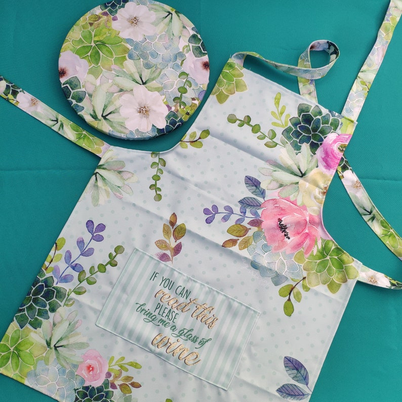 May include: A light blue kitchen apron with a floral print and a pocket that says "If you can read this please bring me a glass of wine".