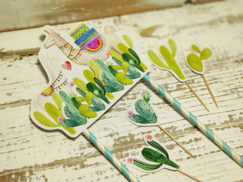 May include: A set of five cake toppers featuring a llama with a colourful saddle and cacti. The llama and cacti are printed on white cardstock and attached to wooden skewers. The skewers are decorated with blue and white striped paper.