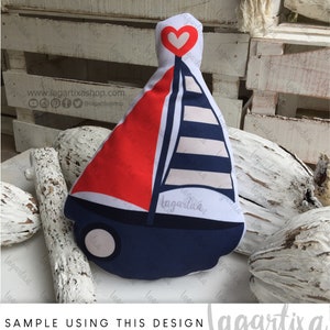 Puede incluir: Una almohada con forma de velero de color rojo, blanco y azul con un coraz&oacute;n blanco en la parte superior. La almohada est&aacute; sobre una superficie de madera blanca con madera flotante.