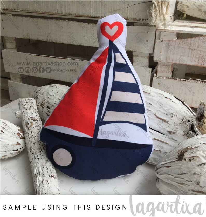 Puede incluir: Una almohada con forma de velero de color rojo, blanco y azul con un coraz&oacute;n blanco en la parte superior. La almohada est&aacute; sobre una superficie de madera blanca con madera flotante.