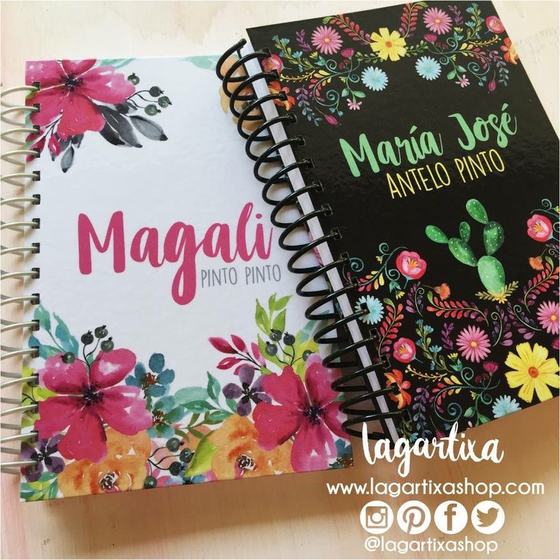May include: Two spiral bound notebooks with floral designs. One notebook is white with pink flowers and the name "Magali Pinto Pinto" printed on the front. The other notebook is black with colorful flowers and the name "Maria Jose Antelo Pinto" printed on the front. Both notebooks have a black spiral binding.