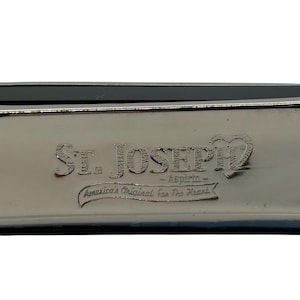 May include: Close-up of a silver metal container with the embossed text "St. Joseph" and "Aspirin." Below the name is the phrase "America's Original For The Heart" in a curved banner.