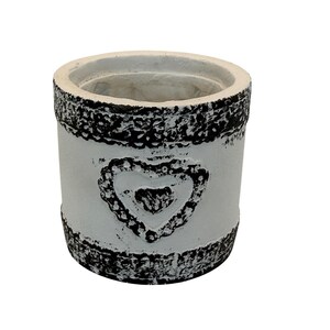 May include: A white cylindrical planter with a textured black rim and base. A heart-shaped design is embossed on the front. The planter is suitable for small plants and adds a decorative touch to any space.