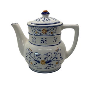 Puede incluir: Tetera de cerámica blanca con tapa, con diseños florales azules y amarillos. La tetera tiene un pico curvo y un asa. La tapa está rematada con un pomo azul y naranja. Diseño Delftware.