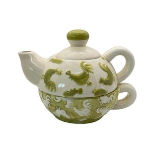 Peut inclure: Service à thé en céramique blanc avec des motifs de coqs verts. La théière est surmontée d'un bouton vert et la tasse est empilée en dessous. L'ensemble présente une anse incurvée.