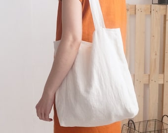 May include: A plain white fabric tote bag with two shoulder straps, worn over the shoulder. The bag is made of a simple, unpatterned textile material and features a rectangular shape with a flat bottom, providing a clean and functional design.