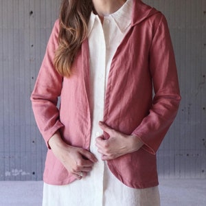 Könnte beinhalten: Eine rosafarbene Jacke aus Leinen mit Kapuze und langen Ärmeln. Die Jacke ist offen und wird über einem weißen Leinenhemd getragen.