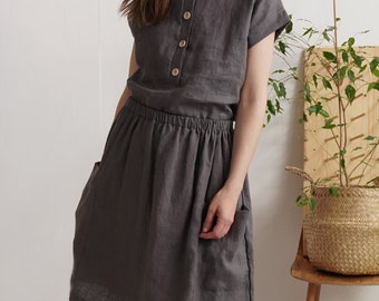 May include: A dark gray dress with short sleeves, a round neckline, and a button-down front featuring four light-colored buttons. The dress has an elasticized waistline and two side pockets, falling to a knee-length hemline.