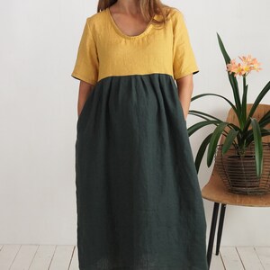 Könnte beinhalten: Zweiteiliges Leinenkleid mit Rundhalsausschnitt und kurzen Ärmeln. Das Oberteil ist senfgelb und der Rock dunkelgrün. Das Kleid hat eine lockere Passform und reicht bis zur Mitte der Wade. Das Model steht vor einer weißen Wand.