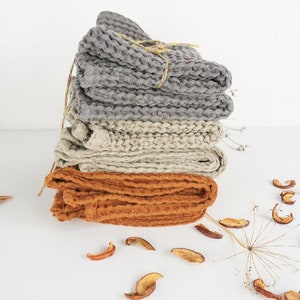 May include: A stack of four waffle weave kitchen towels in gray, beige, and orange. The towels are folded and tied together with twine. The towels are on a white surface with dried apple slices scattered around them.