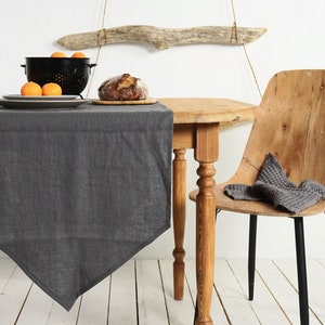 May include: A dark grey linen table runner with a pointed end on a wooden table with a wooden chair. The table runner is on a white wooden surface. There are oranges in a black colander on the table.