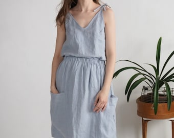 May include: A light blue sleeveless dress featuring a V-neckline with tie-shoulder straps, an elasticized waist, and two large patch pockets on the front of the skirt. The garment is knee-length and has a relaxed, loose-fitting silhouette.