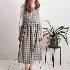 May include: A long, sleeveless apron dress featuring a grey and off-white checkered pattern. The garment has a gathered, elasticized waistline and a full, pleated skirt that falls below the knees. It is worn over a plain, long-sleeved white shirt.