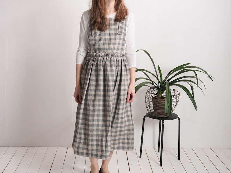 May include: A long, sleeveless apron dress featuring a grey and off-white checkered pattern. The garment has a gathered, elasticized waistline and a full, pleated skirt that falls below the knees. It is worn over a plain, long-sleeved white shirt.