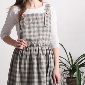May include: A pinafore dress featuring a grey and beige gingham check pattern. The garment has a square neckline, wide shoulder straps secured with light brown wooden buttons, and an elasticated waistband above a gathered, knee-length skirt.