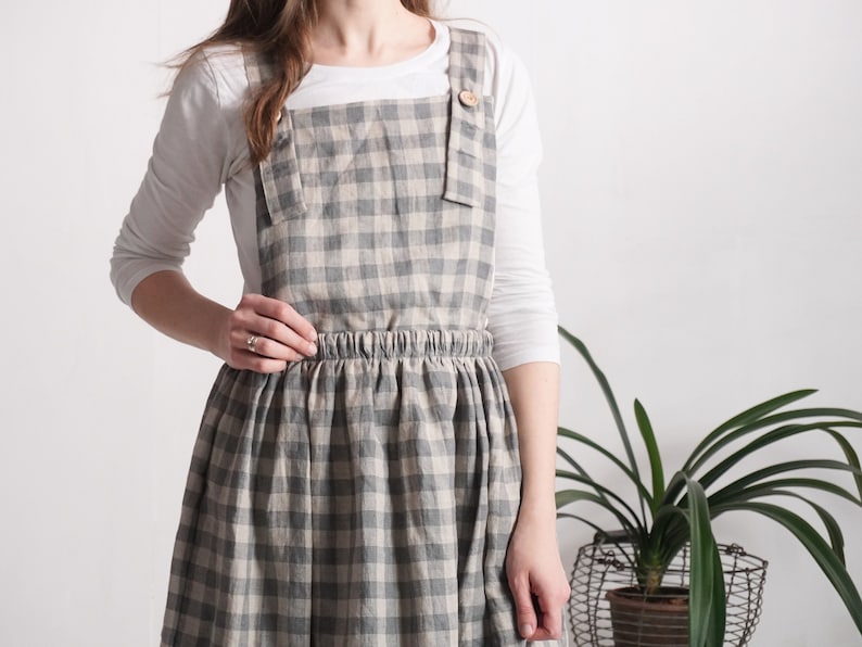 May include: A pinafore dress featuring a grey and beige gingham check pattern. The garment has a square neckline, wide shoulder straps secured with light brown wooden buttons, and an elasticated waistband above a gathered, knee-length skirt.