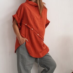 Puede incluir: Una blusa de manga corta color naranja &oacute;xido con cuello alto y una tapeta de botones diagonal en la parte delantera, combinada con pantalones grises holgados con cord&oacute;n en la cintura y un bolsillo visible.