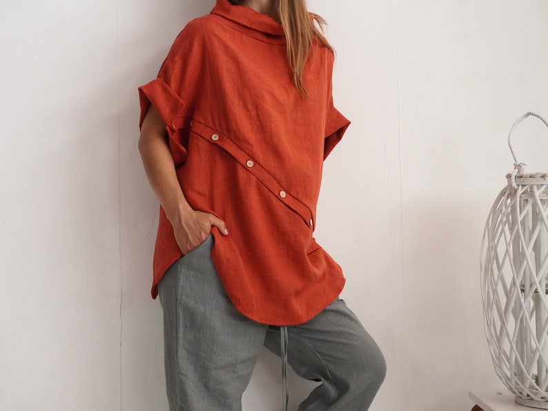 Puede incluir: Una blusa de manga corta color naranja &oacute;xido con cuello alto y una tapeta de botones diagonal en la parte delantera, combinada con pantalones grises holgados con cord&oacute;n en la cintura y un bolsillo visible.