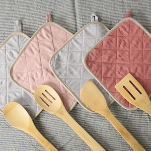 Könnte beinhalten: Vier gequiltete Baumwoll-Topflappen in Grau, Rosa und Rot mit einem beigefarbenen Rand. Drei Küchenutensilien aus Holz liegen auf den Topflappen. Die Utensilien umfassen einen Schöpflöffel, einen Spatel und einen Holzlöffel.