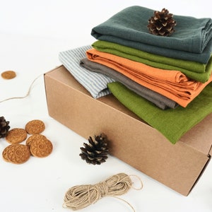 May include: A stack of different colored linen cloths in a brown cardboard box. The box is sitting on a white surface. There are pine cones and small round cookies scattered around the box.